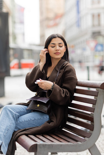 stylish brunette woman with earphone case sitting with closed eyes on bench in prague  - Foto, Bild