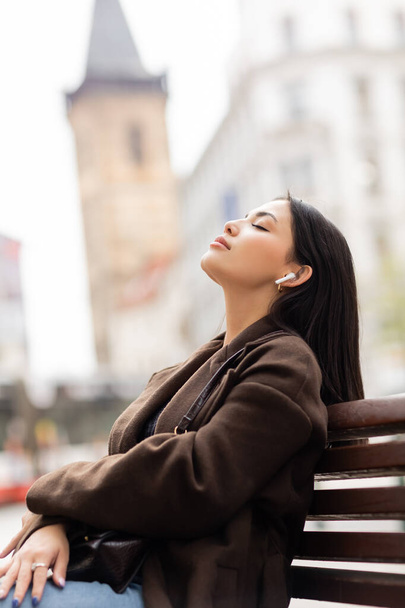 zijaanzicht van de vrouw in de herfst jas ontspannen op de bank tijdens het luisteren muziek in draadloze oortelefoon op straat in Praag - Foto, afbeelding