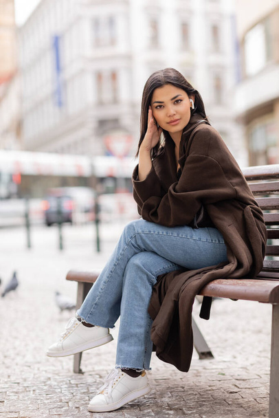 full length of stylish woman sitting on bench in prague and listening music in wireless earphone - 写真・画像