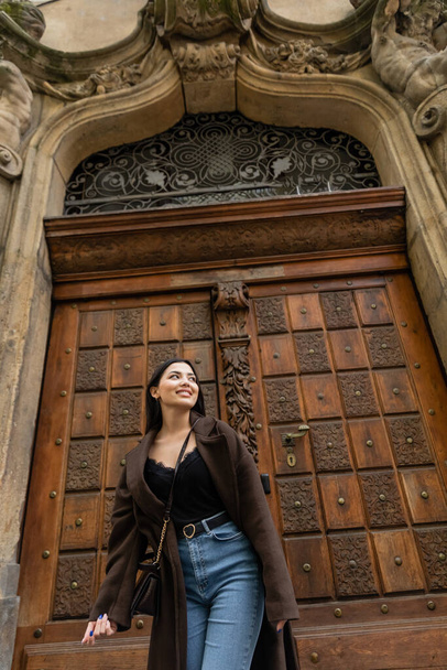 low angle view of brunette woman in jeans and trendy coat smiling near carved wooden door in prague  - Фото, зображення