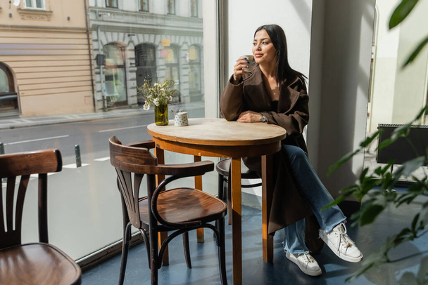 full length of brunette woman looking at city street while sitting in cafe in prague - Fotó, kép