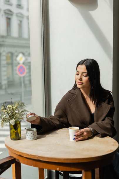 donna bruna con tazza di carta che apre la zuccheriera mentre è seduta vicino alla finestra nel caffè prague - Foto, immagini