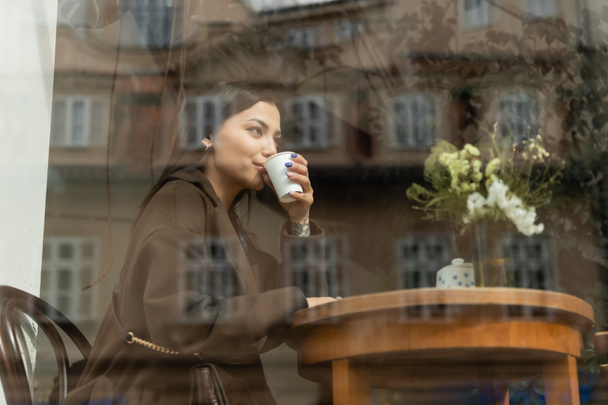 young woman in autumn coat drinking coffee near window in prague cafe - Photo, Image