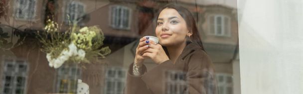 uśmiechnięta kobieta z jednorazowym kubkiem patrząca przez okno w kawiarni Praga, baner - Zdjęcie, obraz