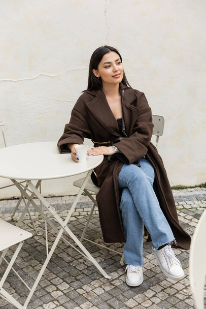 Junge brünette Frau im Mantel hält Pappbecher neben Tisch in einem Café in Prag - Foto, Bild