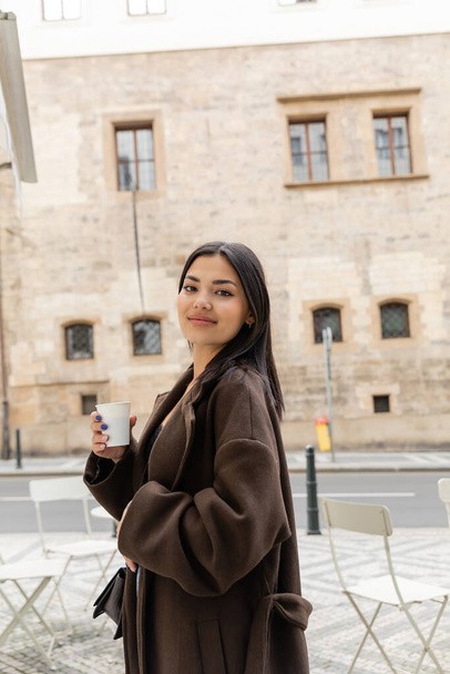 Portrait of young woman in coat holding paper cup and smiling at camera on street in Prague  - Foto, imagen