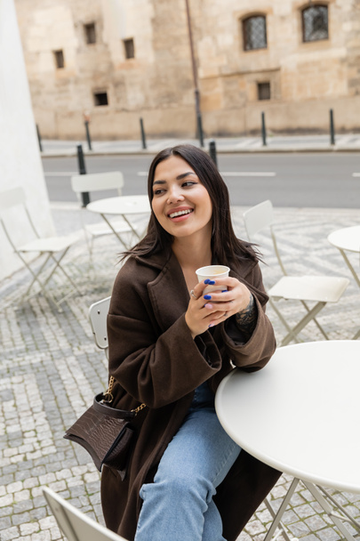 Positivo turista bruna in cappotto che tiene tazza di carta con caffè vicino al tavolo in caffè all'aperto a Praga  - Foto, immagini