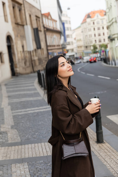 Young brunette woman in coat with handbag holding paper cup near road on urban street in Prague  - Foto, imagen