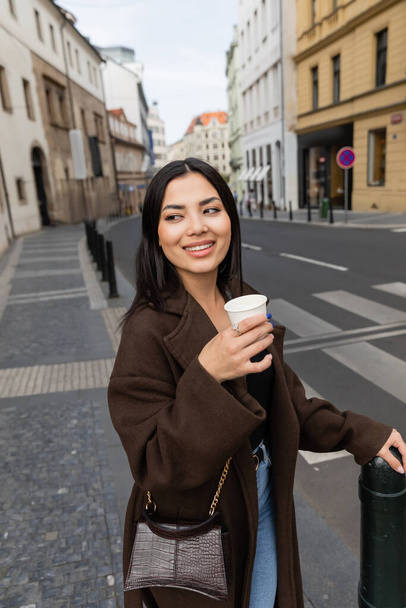 Portrait of stylish young woman holding paper cup with coffee on blurred street in Prague  - Foto, afbeelding