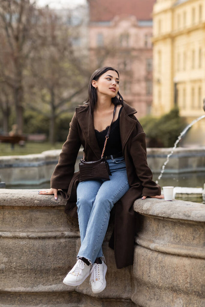 Stylish traveler in coat looking away near paper cup and fountain in Prague  - Foto, Bild