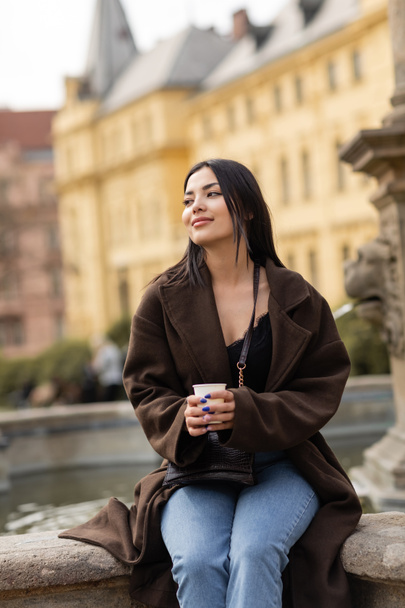 Positive brunette woman in coat holding paper cup near blurred fountain in Charles Square in Prague  - Photo, image