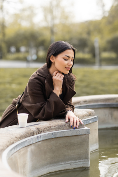 Stylish brunette woman in coat looking at fountain near paper cup on street in Prague  - Photo, Image