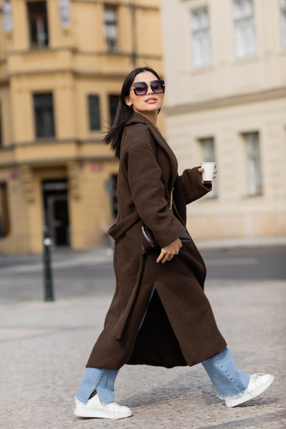 Trendy woman in sunglasses and coat holding takeaway drink on street in Prague  - Fotografie, Obrázek