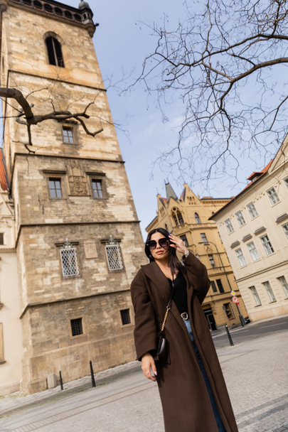 Femme élégante en manteau et lunettes de soleil debout près des bâtiments à Prague  - Photo, image
