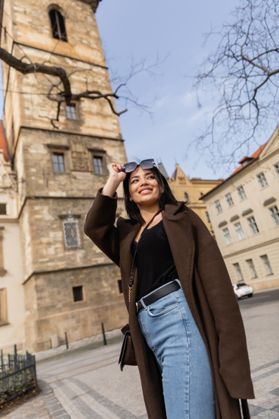 Low angle view of happy traveler in coat holding sunglasses near blurred buildings in Prague  - Foto, Imagem