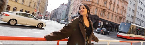 Happy brunette woman standing near road railing on urban street in Prague, banner  - 写真・画像