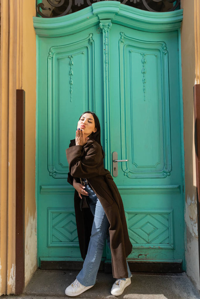 Woman in coat blowing air kiss at camera near turquoise door of building on street  - Фото, изображение
