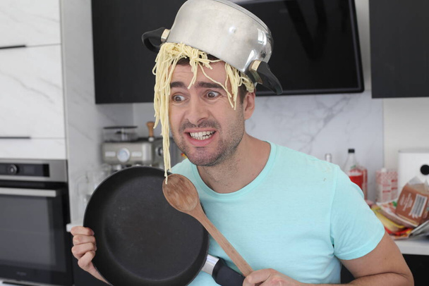portret przystojnego młodzieńca z garnkiem spaghetti na głowie w kuchni - Zdjęcie, obraz