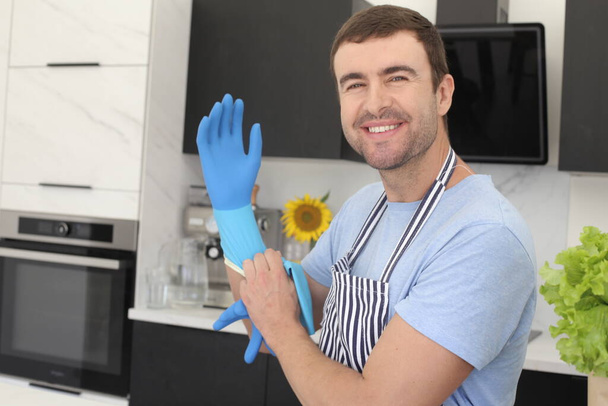 portrait of handsome young man putting on rubber gloves for cleaning at kitchen - Photo, Image