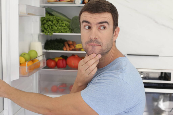 Mutfaktaki buzdolabının önünde duran yakışıklı genç adamın portresi. - Fotoğraf, Görsel