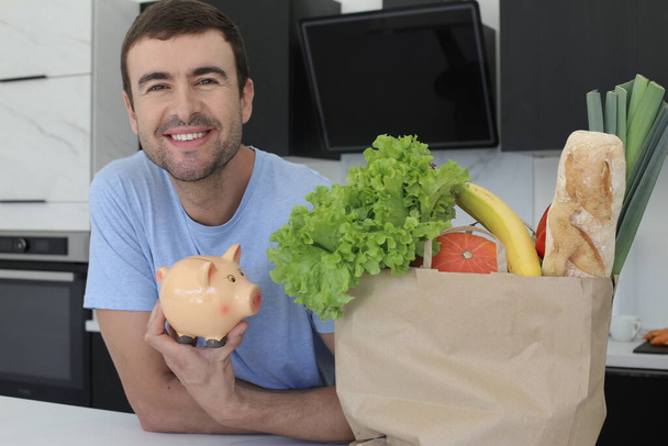 portrait of handsome young man with bag of fresh vegetables holding piggy bank at kitchen - Photo, Image