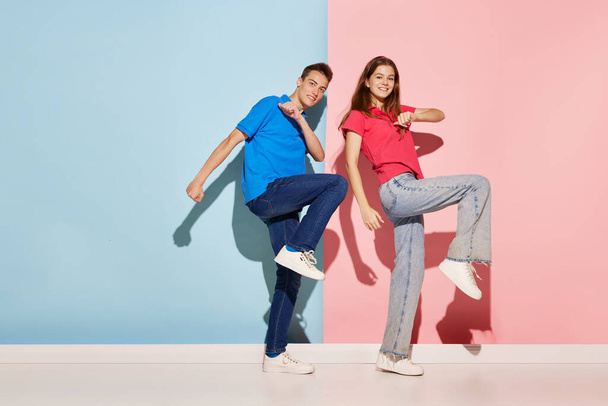 Portrait of young couple, man and woman in casual clothes dancing isolated over blue-pink background. Cheerful. Concept of youth culture, fashion, relationship, emotions. Copy space for ad - Zdjęcie, obraz