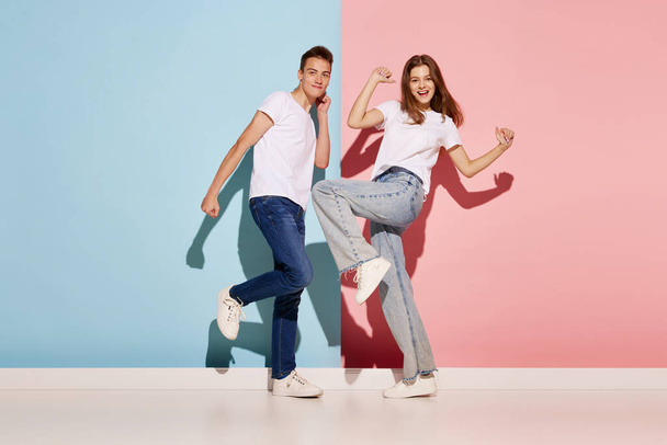 Portrait of young couple, man and woman in casual clothes dancing isolated over blue-pink background. Concept of youth culture, fashion, relationship, emotions. Copy space for ad - Photo, Image