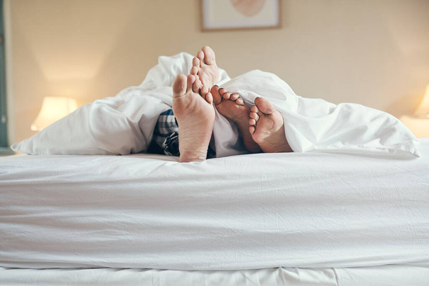 Quedémonos en la cama el resto del día. una irreconocible parejas de pies asomándose debajo de las sábanas mientras yacen en la cama juntos en casa - Foto, imagen