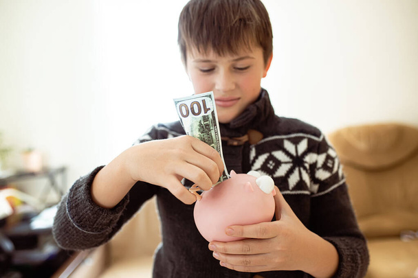 Finger teini laittaa dollareita vaaleanpunaiseen säästöpossuun, lapsi oppii talouslukutaitoa. Nuorten taloudelliset säästöt. Poika, jolla oli rahaa käsissään. - Valokuva, kuva