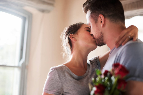 Kissing every moment we can. a young man surprising his girlfriend with a bunch of roses in their bedroom at home - Φωτογραφία, εικόνα