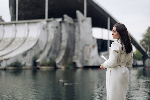 白いコートを着た若い可愛い女性が湖の近くの公園にいる。サイドビュー. - 写真・画像