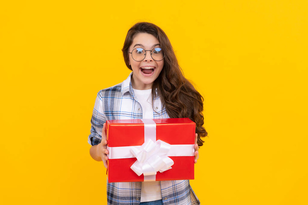 amazed teen girl in glasses with present. studio shot of teen girl hold present. teen girl holding box of present. teen girl with present isolated on yellow background. - Fotó, kép