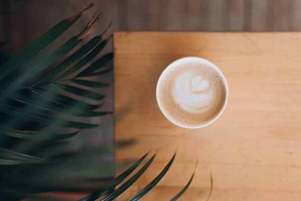 Bovenaanzicht van een papieren kopje koffie met latte kunst op houten tafel. Straatkoffie, bovenaanzicht. Kopkoffie in palmtakken - Foto, afbeelding
