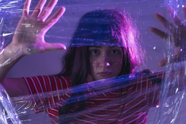 Please, help me - long-haired young Caucasian girl with big dark eyes standing behind the cellophane and touching it with her hands, medium closeup shot with a dark background. High quality photo - Foto, Imagem