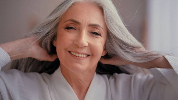 Wunderschöne 60er Jahre alt mittleren Alters reifen kaukasischen Frau Oma Senior ältere Dame 50er Jahre lächelnd in die Kamera zu Hause verwöhnen berühren graue Haare genießen gesunde Frisur Haarpflege-Verfahren - Foto, Bild