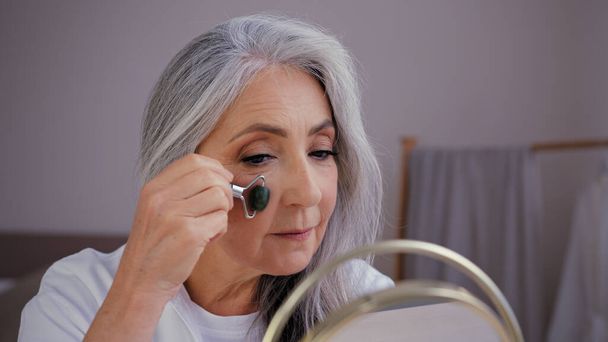 Közelkép öreg ősz hajú kaukázusi nő modell nagymama idős hölgy néz tükörben sima ki ráncok az arcon kvarc kő görgő masszírozó dörzsöli nyirok a bőrön csinál arc fiatalító masszázs - Fotó, kép