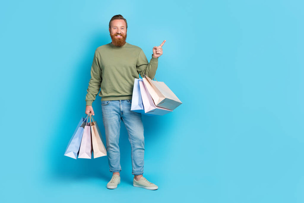 Full size photo of handsome young man hold shopping bags point copyspace dressed stylish khaki look isolated on aquamarine color background. - Valokuva, kuva