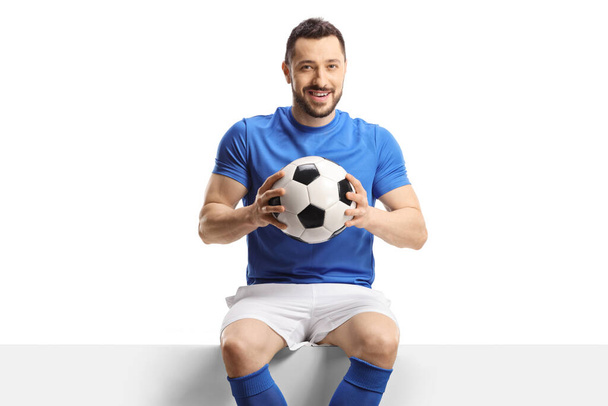 Football player sitting on a blank panel and holding a soccer ball isolated on white background - Foto, Bild