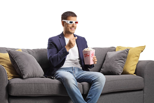 Young man with 3d glasses eating popcorn and sitting on a sofa isolated on white background - Photo, image