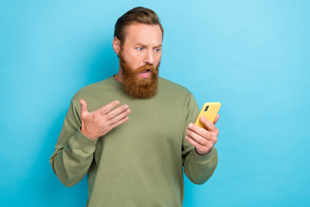 Photo of unsatisfied guy with ginger hairstyle khaki long sleeve look at smartphone read notification isolated on teal color background. - Fotó, kép
