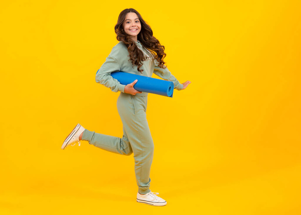 Lunghezza completa di una ragazza adolescente fitness in abbigliamento sportivo tenere tappetino yoga posa su sfondo giallo. Modello fitness bambino che indossa abiti sportivi. Corri e salta. Ragazza nel concetto di sport - Foto, immagini