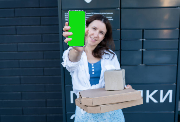 Gelukkige vrouw met mobiele telefoon met groene scherm mockup voor inscriptie en met veel verschillende kartonnen dozen met online bestelling, tevreden met snelle levering, het kopen van producten van hoge kwaliteit - Foto, afbeelding