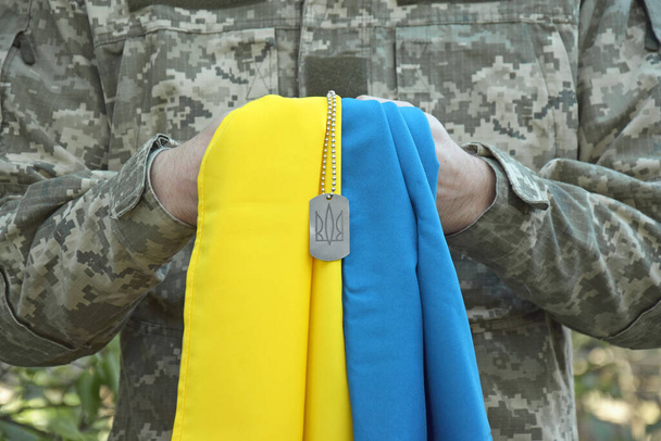 Ένας Ουκρανός στρατιωτικός κρατά την εθνική σημαία στα χέρια του ως σύμβολο του ισχυρού. Πόλεμος στην Ουκρανία. Ημέρα των Κοζάκων και υπερασπιστής της Ουκρανίας. - Φωτογραφία, εικόνα