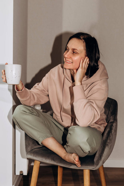 A girl in a pink hoodie sits on a gray chair with a cup of tea in her hands and looks out the window. Front view - Photo, Image