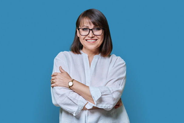 Positive smiling confident middle aged woman looking at camera on blue color studio background. Friendly beautiful mature female with hairstyle makeup wearing glasses, white shirt with crossed arms. - Foto, Imagen