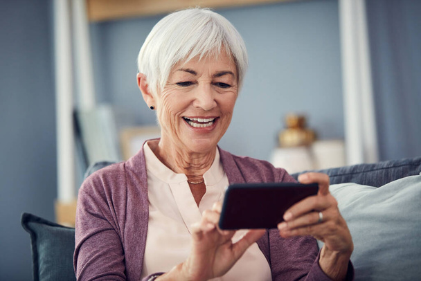 Zwiększę głośność. szczęśliwy starszy kobieta za pomocą smartfona do rozmowy wideo na jej kanapie w domu - Zdjęcie, obraz