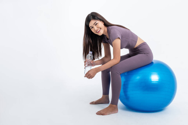 Πορτρέτο εικόνες της Ασίας ελκυστική γυναίκα χαμόγελο και φορώντας ρούχα γυμναστήριο Κρατώντας ένα μπουκάλι νερό και κάθεται στην μπάλα άσκηση, Σε λευκό φόντο, να ασκήσει για την υγεία και την απώλεια βάρους έννοια. - Φωτογραφία, εικόνα