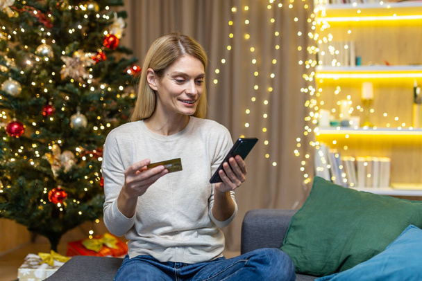 Happy woman alone at home buying Christmas gifts online, housewife sitting on sofa near Christmas tree holding phone and bank credit card, remote shopping for new year. - Foto, Bild