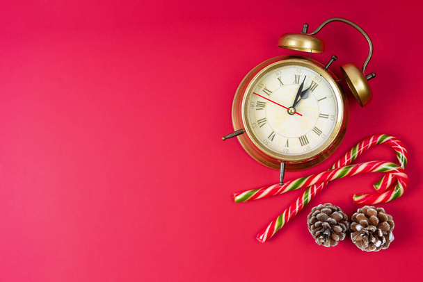 Reloj despertador de bronce, bastón de caramelo y conos de abeto sobre fondo rojo. Las manecillas del reloj muestran doce, medianoche. Vista superior, plano con espacio de copia, banner, encabezado. Navidad y Año Nuevo Fondo - Foto, Imagen