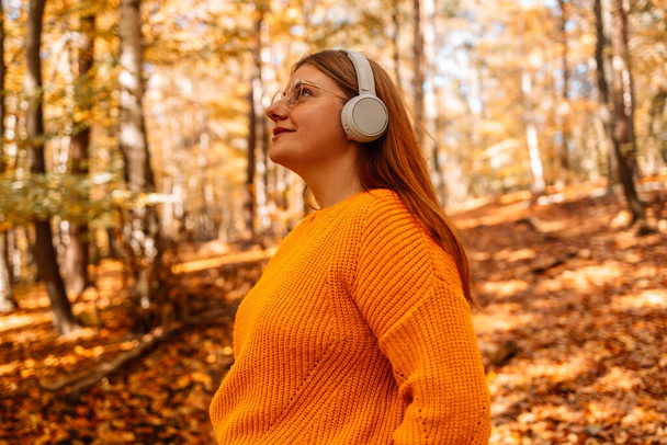 Portret lachende vrouw in een heldere stijlvolle gele trui in een goede bui met behulp van smartphone en het luisteren naar muziek. Meisje met gesloten ogen geniet van haar playlist op smartphone in het herfstbos - Foto, afbeelding